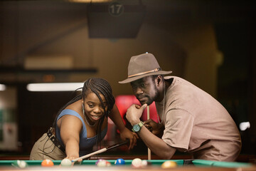Portrait of adult African American couple playing pool together and leaning over table in nightclub
