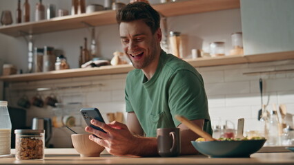 Cheerful man laughing smartphone eating breakfast on kitchen counter closeup