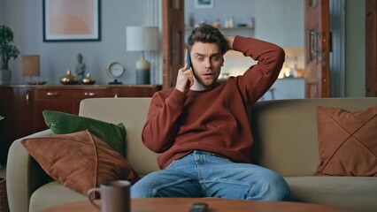 Puzzled man speaking smartphone hearing bad news on couch. Guy feeling headache 