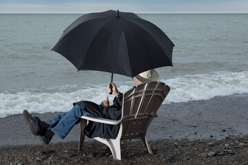 an unidentified person sitting in a beach chair in a raincoat and umbrella looking at dull waves...