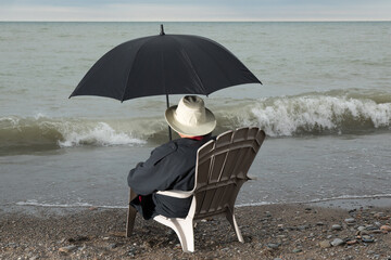 the back of an unidentified person sitting in a beach chair in a raincoat and umbrella looking at...