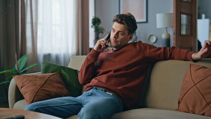 Bored guy calling cellphone relaxing at couch. Serious man talking smartphone