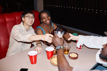 Multiethnic group of friends clinking beer bottles and cheering sitting at diner table together - 782463237