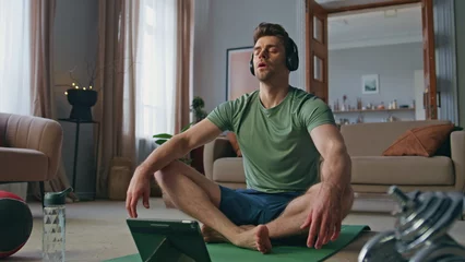 Fotobehang Serene athlete meditating headphones at apartment. Calm fit man sitting in lotus © stockbusters