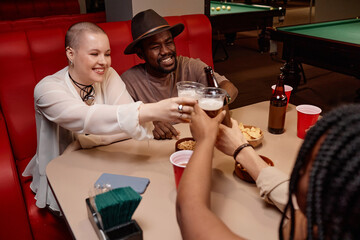 Multiethnic group of friends clinking beer glasses and cheering sitting at diner table