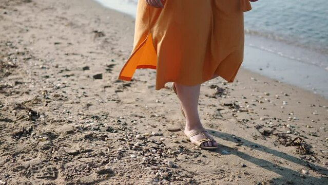 Legs of white woman in dress and sandals walk along sandy shore along sea closeup. Waves roll onto beach and wind flutters hem of orange dress. Stroll on early sunny morning.