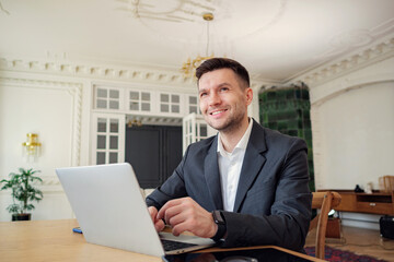 Professional businessman in a classic suit smiles while working on a laptop in a bright, elegant...