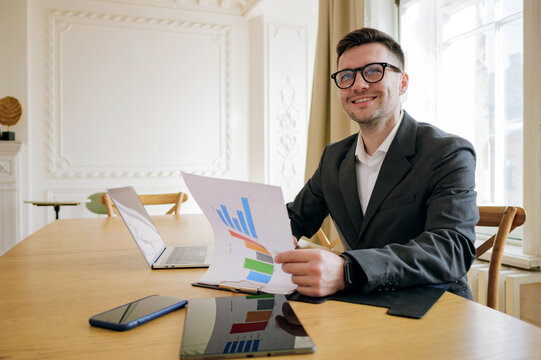 Analytical businessman examines graphs with a confident smile, amidst a serene office with tech aids at his disposal.