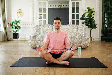A tranquil man in a pink shirt sits cross-legged, practicing meditation, eyes closed, on a mat in...