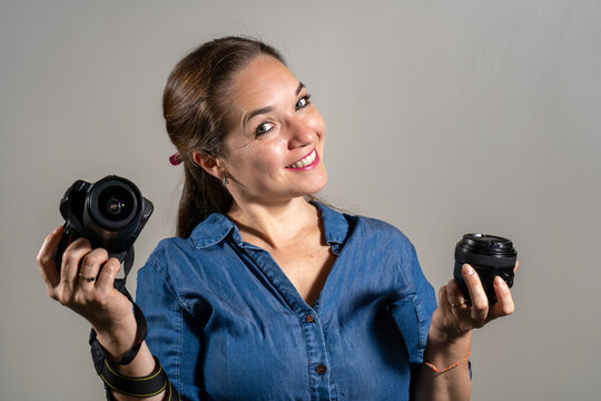 Mujer sosteniendo su cámara digital en una mano, y un lente de fotografía en la otra, mientras sonrie al espectador