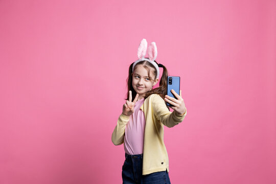 Joyful happy child taking pictures and showing peace sign on phone, feeling cheerful and confident about fun photos for easter celebration. Little schoolgirl with pigtails takes photographs.