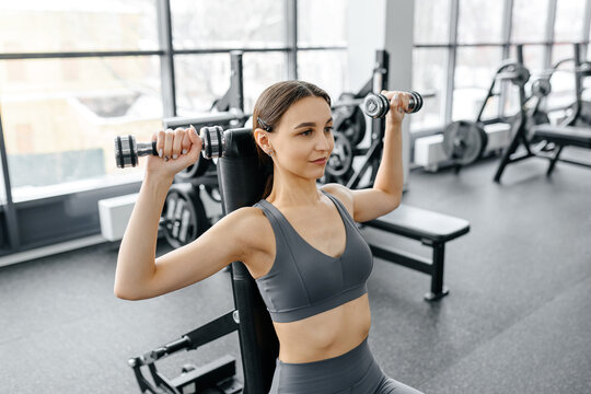 young athletic caucasian woman trains in fitness gym, doing dumbbell press up on bench, brunette girl in grey top and leggings, healthy lifestyle concept