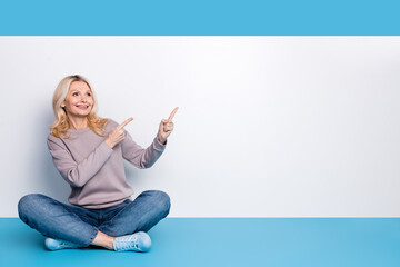 Full body photo of senior lady sit floor crossed legs point empty space dressed stylish gray garment isolated on blue color background