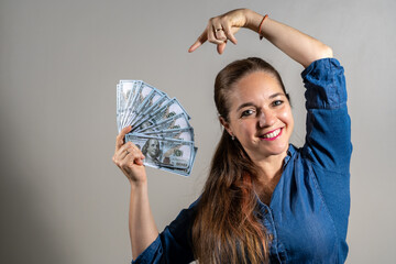 Mujer sosteniendo un monton de dolares mientras señala el dinero y sonrie a cámara - obrazy, fototapety, plakaty