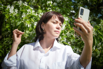 Brunette girl Using Smartphone in Blossoming Orchard in Springtime. Middle aged Woman taking selfie by phone among spring blossoms of apple trees