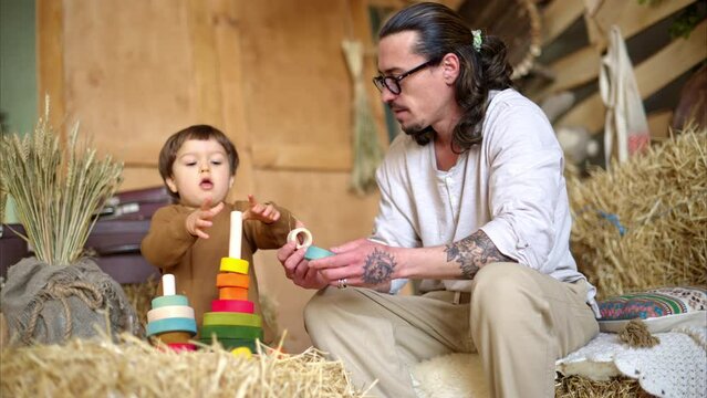 Father playing with his son with colourful, ecological wooden toys in a barn, near square hay bales