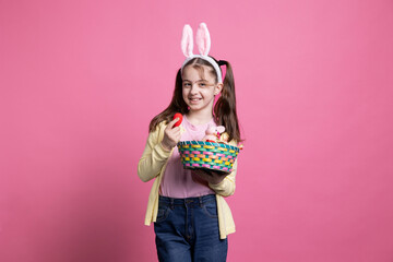 Adorable joyful child holding colorful eggs and a stuffed rabbit on an easter arrangement, posing in pink studio. Young smiling girl with bunny ears holding a basket with lovely spring ornaments.