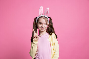 Obraz na płótnie Canvas Smiling little child showing peace sign in front of camera, feeling excited about gifts and easter festivity. Young girl with bunny ears posing in studio, being happy and positive about spring.