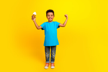Full length photo of cheerful child with curly hair dressed blue t-shirt hold ice cream raising fist up isolated on yellow background