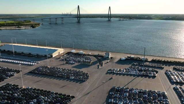 New cars parked in vast lots with Arthur Ravenel Jr. Bridge in the background at sunrise.