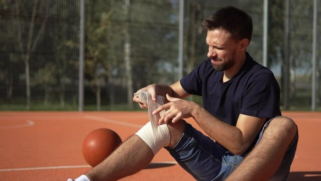 Caucasian man sits on basketball court and bandages knee with elastic bandage after injury. Suffering and pain due to dislocation and fixation of joint after sports activity on outdoor sports field.