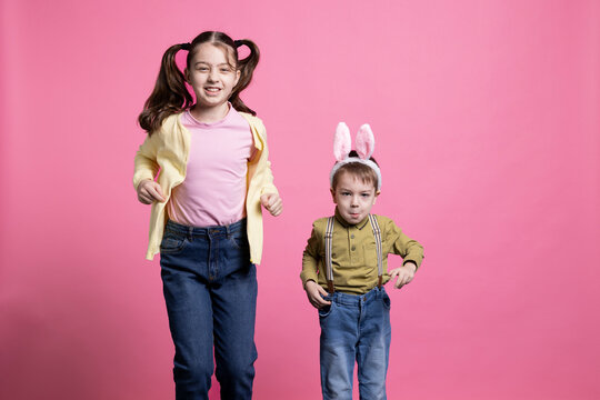 Brother with bunny ears and sister jumping around in the studio, little children being happy about easter time celebration. Adorable siblings bouncing like rabbits against pink background.