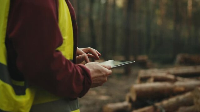 At a work site among the forests, a lumberjack engineer stands, carefully studying the data on his tablet. Dressed in a safety helmet and a colorful vest, he monitors the cutting process