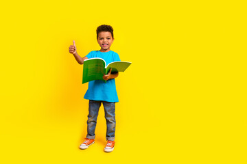 Full body photo of satisfied little child dressed blue t-shirt jeans hold book showing thumb up isolated on vibrant yellow background