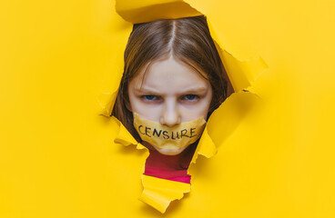 A child with his mouth taped shut peeks through a torn hole in yellow paper.The inscription on the tape is 