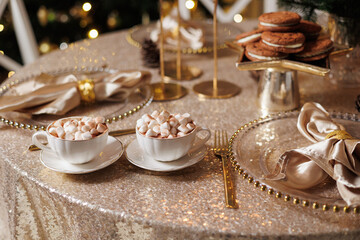 Christmas or New Years festive party luxury table setting. Glittering tablecloth, cups with cacao and marshmallow, chocolate cookies, glasses plates, napkins, golden cutlery, candlestick, garlands