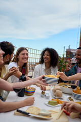 Vertical. Group of multi-ethnic young friends enjoying meal gathered on rooftop. Happy millennial...