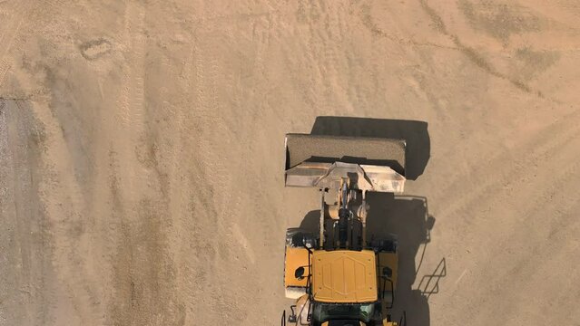 Aerial shot - Drone following an excavator with fully loaded arm driving on gravel road to a special machine, into which he dumps the gravel.