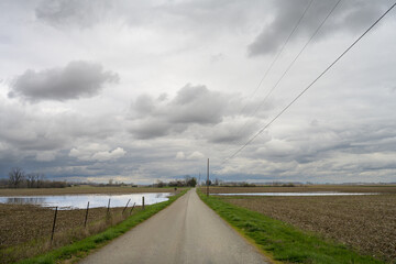 A country road on a moody, rainy, stormy day. The sky has dramatic clouds and the fields have pools of water from recent rains. The road extends into the distance with telephone poles and an old fence - Powered by Adobe