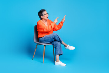 Full body photo of lovely senior lady sit chair look point empty space dressed stylish orange garment isolated on blue color background