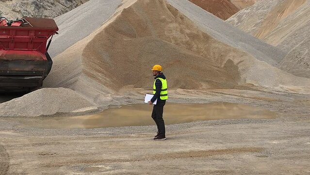 Quarry supervisor walking around the quarry in reflective vest and protective helmet with notebook in his arm. Walking past heavy machinery and large piles of gravel.