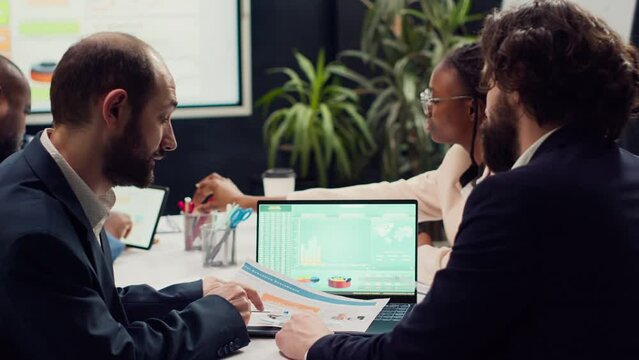 Business partners examining sales infographics in a boardroom, reviewing market trends and competitive analysis. Specialists share ideas for new products, ensuring company success. Camera A.
