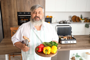 Mature man with board of fresh vegetables and tasty sausage cooked on modern electric grill at table in kitchen