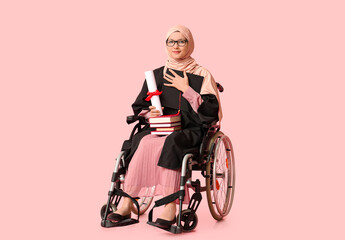 Beautiful female Muslim graduate student in wheelchair with diploma, graduation cap and books on pink background