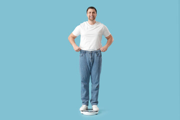 Fototapeta na wymiar Young man in loose jeans measuring weight on scales against blue background