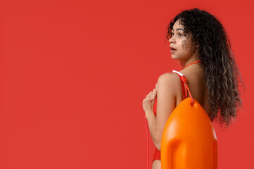 Beautiful young African-American female lifeguard with rescue buoy on red background