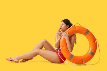 Beautiful young African-American female lifeguard with ring buoy whistling on yellow background