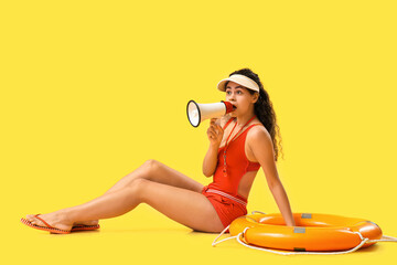 Beautiful young shocked African-American female lifeguard with ring buoy and megaphone on yellow...