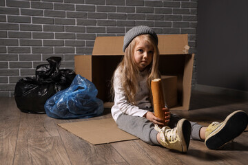 Homeless little girl eating bread near trash against grey brick wall - obrazy, fototapety, plakaty