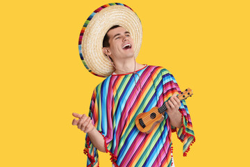Young man in Mexican sombrero hat and poncho playing ukulele on yellow background - obrazy, fototapety, plakaty