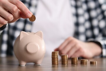 Financial savings. Man putting coin into piggy bank at wooden table, closeup - obrazy, fototapety, plakaty