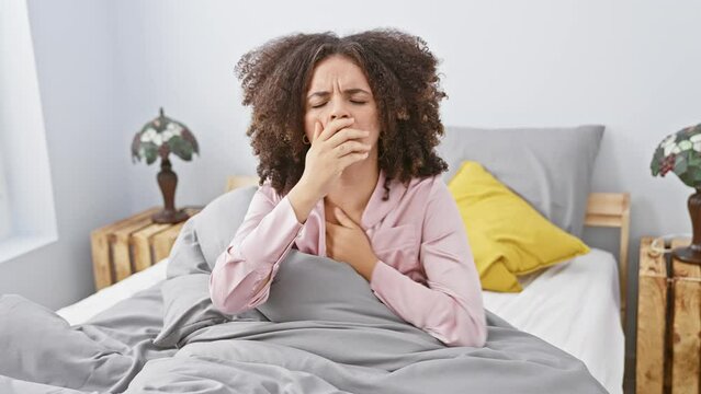 A young hispanic woman with curly hair appears ill, sneezing and clutching chest, in a bedroom setting.