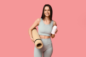 Young sporty woman with water bottle and yoga mat on pink background