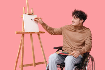Young male artist on wheelchair with brush and palette near canvas on pink background