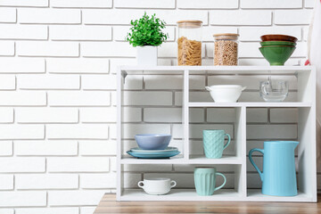 Shelving unit with set of clean tableware and cups near white brick wall