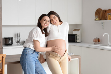 Young lesbian woman touching belly of her pregnant wife in kitchen